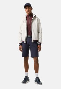 Leichte, weiße Reisverschlussjacke mit kastanienbraunen Akzenten, über einem kastanienbraunen Langarmshirt und navy Shorts, kombiniert mit dunklen Turnschuhen.