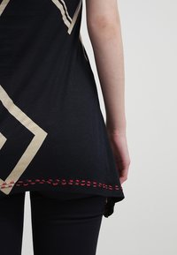 Black top with beige geometric pattern and red stitched hem. Cotton material, flowy shape, features a rounded bottom.