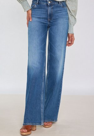 Femme portant un jean bleu taille haute à jambes larges, un chemisier vert clair à motifs floraux et des sandales à plateforme marron à talons blocs.