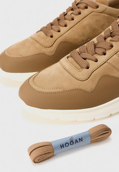 Un paio di scarpe da ginnastica marroni con dettagli testurizzati, suole bianche spesse e un paio di lacci di ricambio abbinati etichettati "Hogan" posti sotto le scarpe.