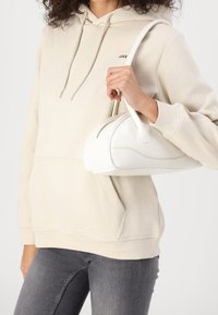 Borsa in pelle bianca con forma arrotondata, dettaglio di cucitura curvata e manico superiore, abbinata a un hoodie beige e jeans grigi.