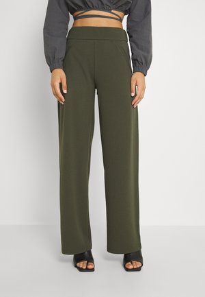 Femme portant un pantalon large taille haute vert olive, un top court noir à manches longues, et des chaussures à talons ouverts noires.