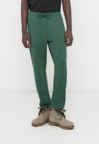 Πράσινο παντελόνι φόρμας με σούρα στη μέση, εφαρμοστή γραμμή και απαλό ύφασμα, συνδυασμένο με καφέ μπότες δέτου τύπου με ανθεκτικό σχέδιο.