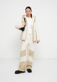 Doudoune claire avec capuche, blocs de couleur crème et beige ; pantalon assorti avec poches et texture à motifs ; sac à main noir et bottes.