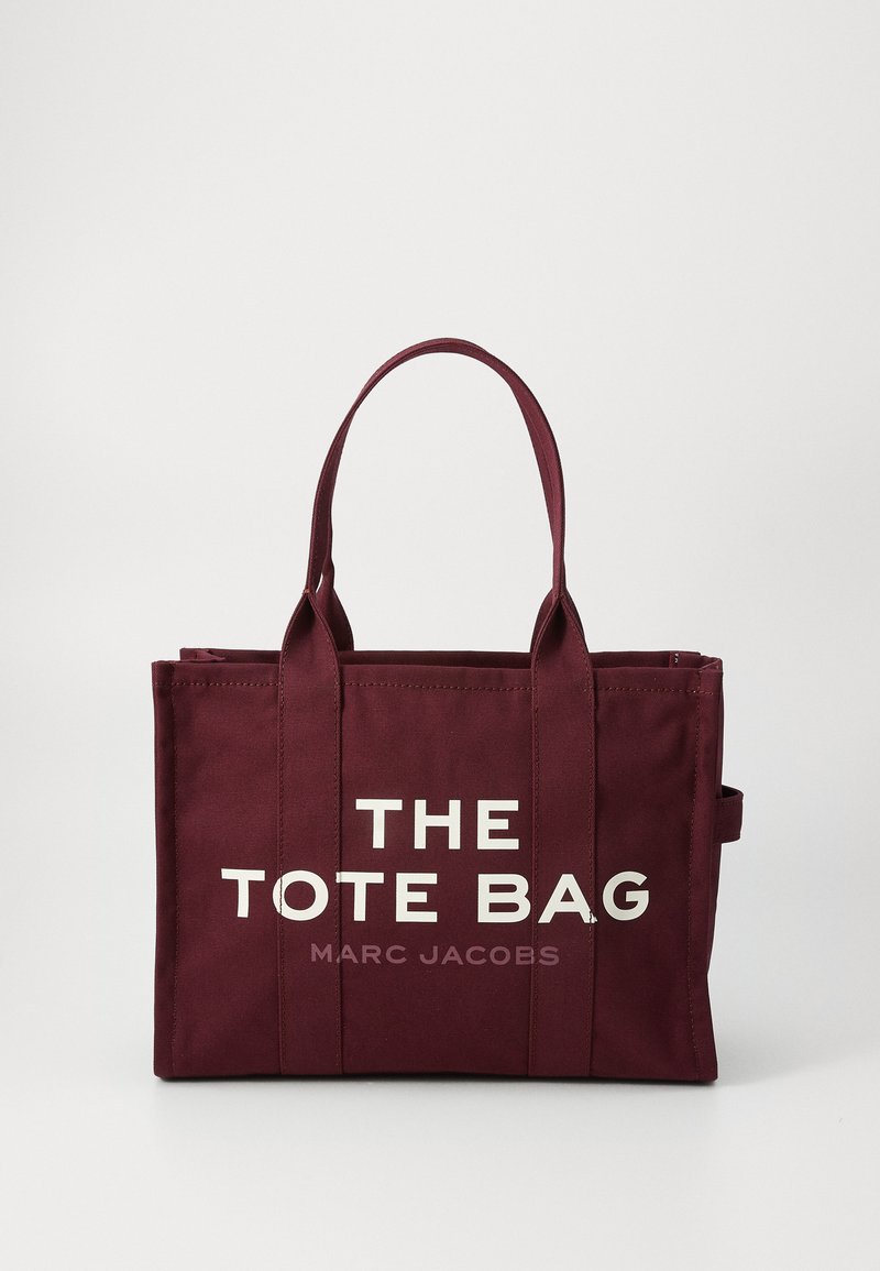 Borsa a tracolla in tela bordeaux con due manici, caratterizzata da una grande scritta bianca che recita "THE TOTE BAG" e una scritta più piccola "MARC JACOBS" sotto.