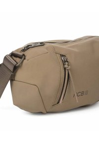 Beige stoffen crossbodytas met een afgeronde vorm, voorzien van een voorvak met rits, een gladde textuur en een logodetail aan de zijkant.