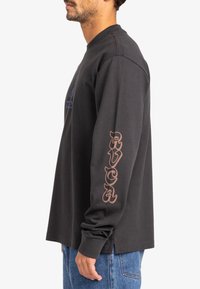 Zwarte katoenen t-shirt met lange mouwen, voorzien van een blauwe afbeelding op de voorkant en bruine "RVCA" tekst op de mouw. Het shirt heeft een relaxte fit en zijopeningen.