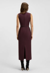 Midi-kjole i mørk bordeaux, tætsiddende design, ærmeløs, med en slids bagpå og sømløs konstruktion. Kombineret med knælange sorte støvler.