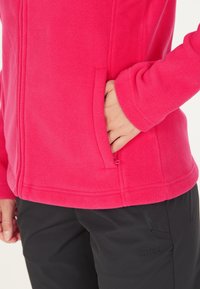 Fuchsia fleecejakke med en lynlås lomme og en glat tekstur. Har en tætsiddende design med syninger og bæres med sorte bukser.