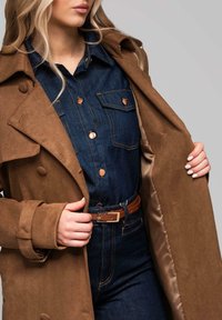 Gabardina de ante marrón sobre una camisa oscura de mezclilla con botones de cobre. La gabardina tiene una cintura con cinturón y dos grandes bolsillos delanteros.