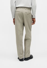 Άνδρας που φοράει ανοιχτόγκρι παντελόνι φόρμας με κουμπωμένες πίσω τσέπες, συνδυασμένο με μαύρα παπούτσια φόρμας και ανοιχτόγκρι πουκάμισο, όρθιος με την πλάτη.