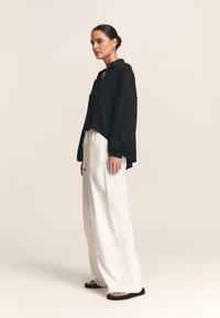 Črna srajca s gumbi, dolgimi rokavi in ukrivljenim robom, kombinirana z belimi širokimi hlačami in črnimi natikači. Gladek tekstil.