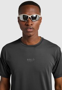Svart kortärmad träningsskjorta med en reflexbelagd "HALO" logotyp. Modellen bär ljusa insvepande solglasögon med spegelglas.