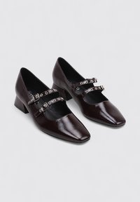 Zapatos de tacón bloque de cuero charol marrón oscuro con punta afilada y detalles de doble correa con herrajes en tono plateado.