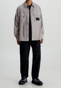 Grå rutiga skjorta med två bröstfickor och en svart etikett, ihop med svarta byxor och svarta sneakers med vita accenter.