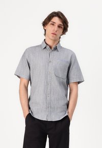 Chemise à manches courtes en tissu gris clair avec une texture subtile, dotée d'une poche poitrine et de détails de couture contrastés en bleu.