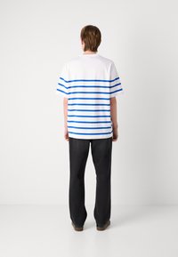 Weißes kurzärmliges Hemd mit blauen horizontalen Streifen, lockere Passform, Rückansicht. Kombiniert mit schwarzen Hosen und braunen Schuhen. Einfaches Design.