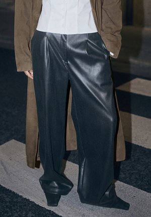 Asmuo, vilkintis laisvas juodas tekstūruotas odines kelnes, baltą marškinį ir rudą suaugusią paltą, stovi perėjimo zonoje naktį.