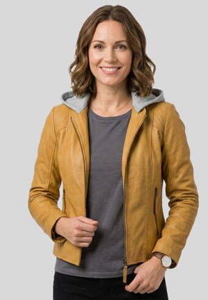 Lächelnde Frau mit welligem braunem Haar trägt eine senffarbene Lederjacke über einem grauen Shirt und steht vor einem schlichten hellen Hintergrund.