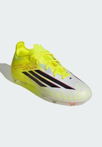 Κίτρινο ποδοσφαιρικό παπούτσι με υφασμένο λευκό επάνω μέρος, διακοσμημένο με μαύρες ρίγες και κόκκινο λογότυπο. Διαθέτει κλείσιμο με κορδόνια και καρφιά για πρόσφυση.