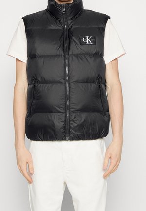 Czarna pikowana kamizelka bez rękawów z przednim zamkiem błyskawicznym, kieszeniami na zamek i naszywką z logo Calvin Klein Jeans, noszona na białej koszulce z krótkim rękawem.