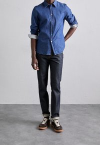 Denim-painikkeellinen paita keskisinisessä sinisessä, jossa on kaulus, raidalliset kalvosimet ja värikkäät napit, yhdistetty tummiin farkkuihin ja ruskeisiin mokkanahkaisiin tennareihin.