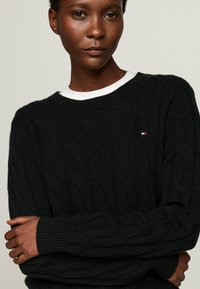 Pull en maille noire avec un col rond et des poignets côtelés, présentant un petit logo sur la poitrine. Tissu lisse et texturé.