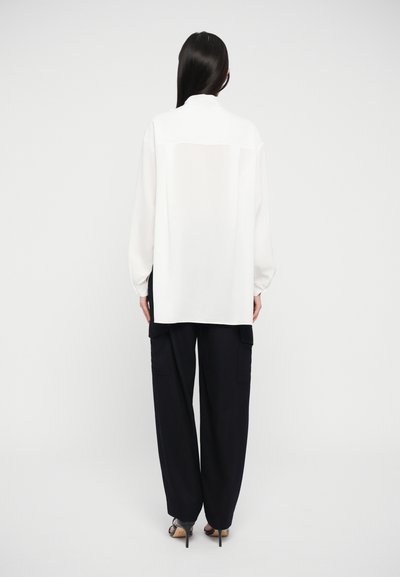 White long-sleeve blouse with a relaxed fit, featuring a hidden button placket and a yoke detail at the back. Paired with navy wide-leg trousers.