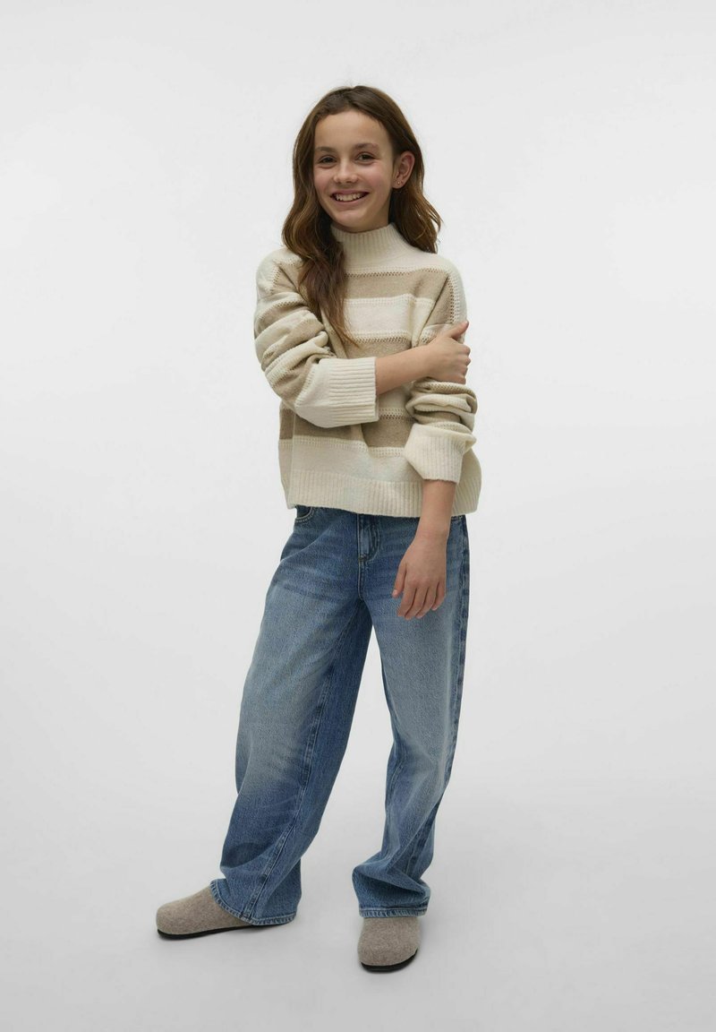 Pleteni krem pulover z bež črtami, kroja z okroglim izrezom in z ribastimi manšetami, v kombinaciji s širokimi svetlo modrimi jeans hlačami in sivimi natikači.