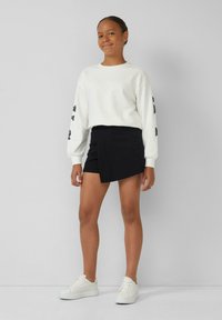 Une jeune personne se tient souriante, vêtue d'un sweat-shirt blanc, d'une jupe asymétrique noire et de baskets blanches, devant un fond gris uni.