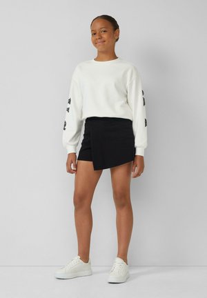 Une jeune personne se tient souriante, vêtue d'un sweat-shirt blanc, d'une jupe asymétrique noire et de baskets blanches, devant un fond gris uni.