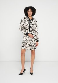 Φόρεμα από κρεμ ύφασμα με μαύρες αφηρημένες ρίγες, μακριά μανίκια, κουμπιά στο μπροστινό μέρος και εφαρμοστή μέση. Συνδυάζεται με μαύρα ψηλοτάκουνα παπούτσια.