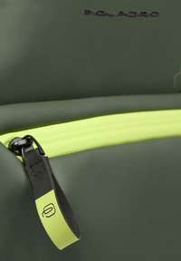 Donkergroen rugzakmateriaal met een opvallende gele rits. De ritssluiting heeft een zwart accent met een logo.