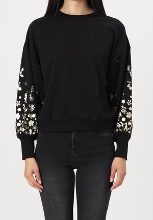 Femme portant un sweat-shirt noir à manches longues avec des motifs floraux métalliques sur les manches et un jean noir, debout devant un fond blanc.