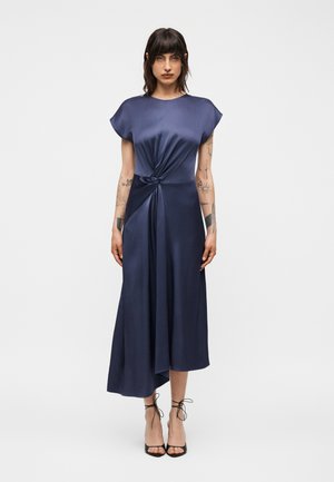 Kvinde med mørkt hår og tatoveringer iført en mørkeblå satin kjole med knudedetalje og sorte remsko, stående mod en enkel hvid baggrund.