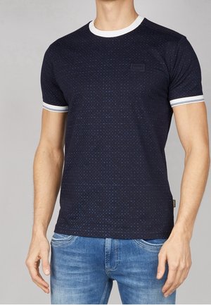 Mężczyzna ubrany w granatowy, wzorzysty t-shirt z krótkim rękawem, z białym kołnierzykiem i mankietami na rękawach, w zestawie z niebieskimi jeansami.