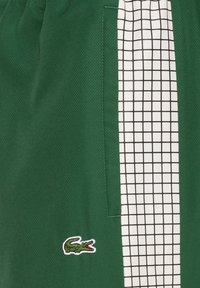 Pantalones cortos verdes con tejido texturizado, panel lateral con patrón de cuadrícula negro y un logotipo bordado en blanco de un cocodrilo.