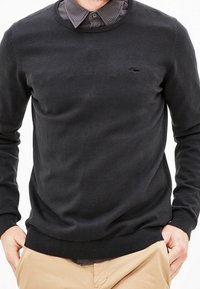 Schwarzer Strickpullover mit Rundhalsausschnitt, gerippten Bündchen und Saum. Über einem karierten Hemd mit verstecktem Kragen getragen. Dezentes Logo-Detail.
