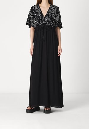 Žena v černých maxi šatech s bílou květinovou výšivkou na krátkých rukávech a horní části šatů, doplněná černými sandály na platformě.