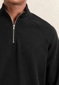Mann trägt einen schwarzen Pullover mit Viertelreißverschluss und silbernem Reißverschluss, von der Mitte der Brust bis zum Hals vor einem neutralen Hintergrund dargestellt.
