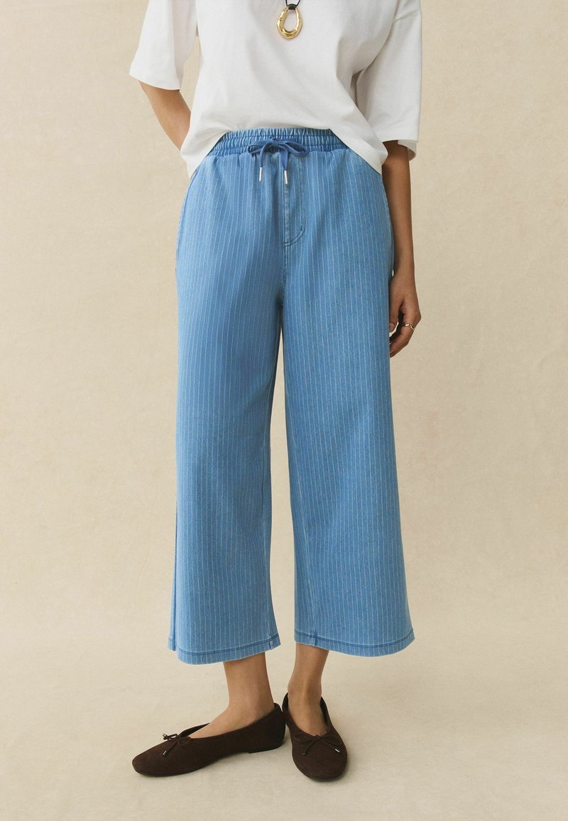 Personne portant un pantalon large raccourci à fines rayures bleu clair, une chemise blanche à manches courtes, des ballerines noires et un collier pendentif en or, debout.