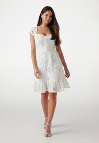 Abito bianco in pizzo con maniche corte, un corpetto aderente e un orlo svasato. Presenta motivi intricati e una gonna arricciata. Tacchi argentati.