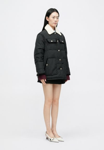 Sort pufferjakke med faux fur krave, der har trykknapper og forlommer. Modellen bærer en kort nederdel og hvide hæle.