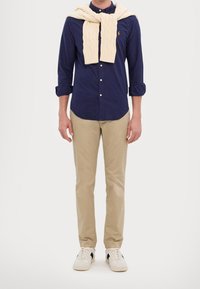 Navy button-up shirt, cream cable-knit sweater draped over shoulders, beige chinos, and white sneakers with black accents.