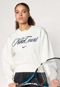 Λευκή φούτερ μπλούζα με ναυτική «NikeCourt» κέντημα και ριμπέτ μανίκια, συνδυασμένη με σκούρα παντελόνια και ένα ρακέτα τένις στο προσκήνιο.