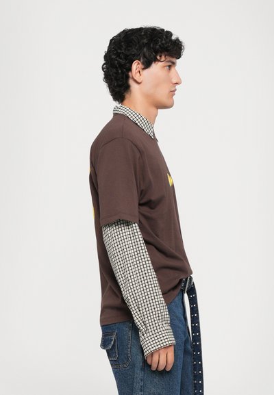 Mladý muž s kučeravými vlasmi, oblečený v hnedej vrstvenej tričku cez vzorovanú košeľu s dlhými rukávmi a modrými džínsami, stojí v profile na bielom pozadí.