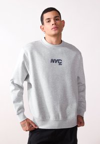 Felpa grigia chiara con maniche lunghe, scollatura rotonda e la scritta "NYC 86" stampata in blu scuro sul petto. Tessuto morbido, vestibilità comoda.