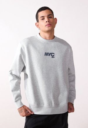 Svijetlo siva dukserica s dugim rukavima, okruglim izrezom i otiskom "NYC 86" u tamnoplavoj boji na prsima. Mekana tekstura, opušteni kroj.