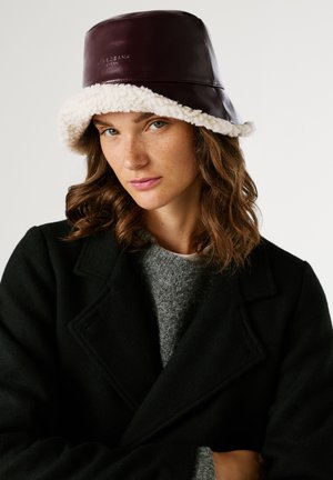 Femme avec des cheveux bruns ondulés portant un chapeau en shearling sombre, un manteau noir et un pull gris, regardant l'appareil photo sur fond blanc.