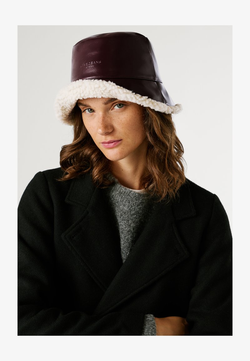 Donna con capelli castani mossi che indossa un cappello scuro in pelliccia, un cappotto nero e un maglione grigio, che guarda la camera su uno sfondo bianco.